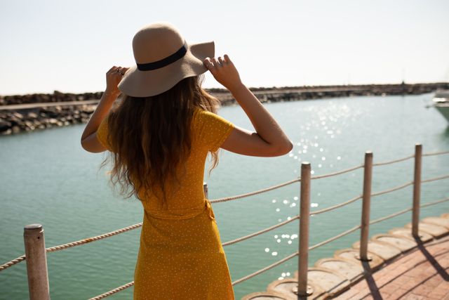 Rear view of teenage Caucasian girl, wearing a straw hat, enjoying her time on a promenade by the sea, on a sunny day, wearing a sun hat admiring the view.