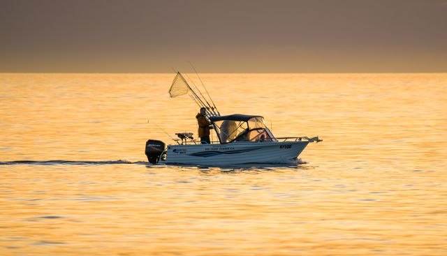 Man standing on small fishing boat during sunset, holding fishing rod and net. Golden sky reflecting on calm ocean water creates peaceful atmosphere. Ideal for depicting outdoor hobbies, leisure activities, solitude, and nature scenes.