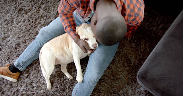 Man sitting on carpet petting dog in living room, highlighting companionship and comfort in a home environment. Perfect for themes related to pets, relaxation, friendship, and home lifestyle.