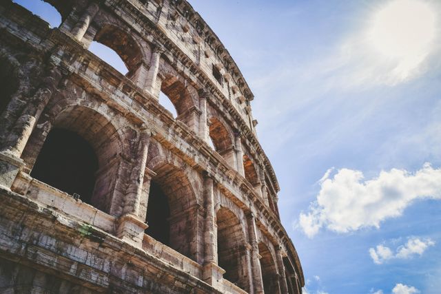 Photo showing the ancient Roman Colosseum in Rome, Italy with its iconic arches and historical architecture set against a clear blue sky and bright sun. Perfect for articles about Roman history, architecture tourism, travel destinations, and educational content on historical landmarks.