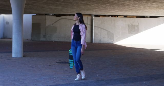 Happy biracial woman walking with skateboard in underground parking, and looking ahead on sunny day. Free time, lifestyle, city life, sport and hobby, unaltered.