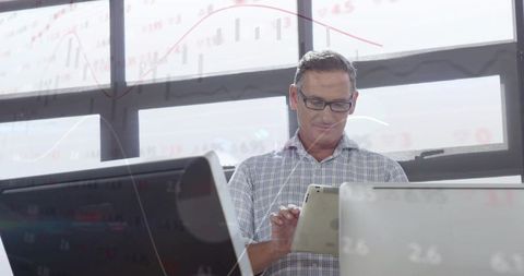 Mature professional reviewing tablet while surrounded by laptops and financial charts