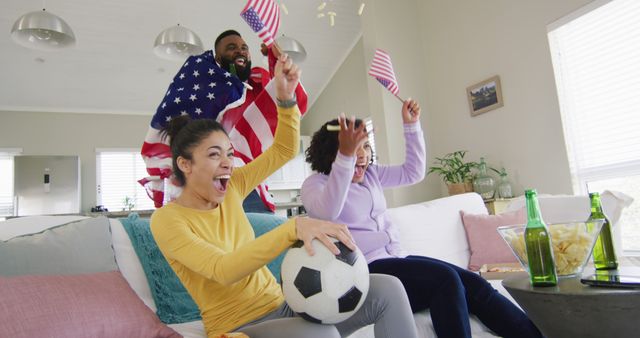 Group of friends seen cheering and celebrating a soccer match in their living room. One person is enthusiastically waving an American flag while the others are holding mini American flags. The vibe is joyous and full of team spirit, ideal for setting a scene of fan engagement, sports enthusiasm, or national support. Perfect for stock images relating to sports events, fandom communities, and home gatherings.
