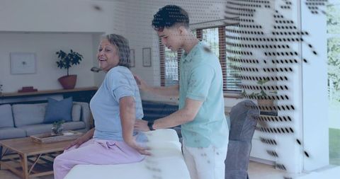 Young caregiver assisting senior woman sitting on bed in sunlit living room, home care support