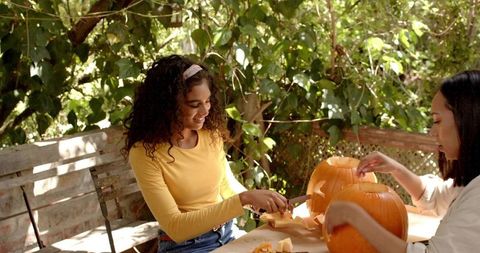Friends carving pumpkins on sunny backyard patio, autumn pumpkin carving activity