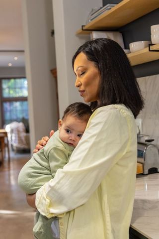 Mother holding infant close in sunlit kitchen, cradling newborn for tender bonding