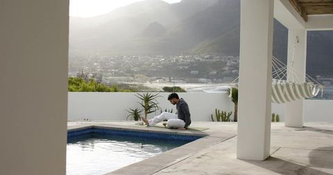 Indian man using laptop by pool on coastal villa terrace, hammock and minimalist retreat