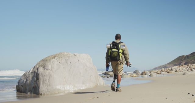Rear view of biracial man with prosthetic leg trekking with backpack on a beach by the sea. Long distance walking, fitness, challenge, disability, nature and healthy outdoor lifestyle.