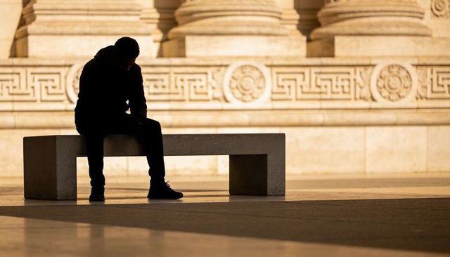 Sitting shadowed man leaning forward on bench in neoclassical forecourt at golden hour