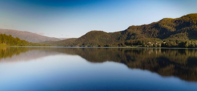 Peaceful mountain lake reflecting surrounding hills and forest under a clear blue sky. Ideal for use in nature calendars, travel brochures, and environmental campaigns promoting the beauty of untouched landscapes.