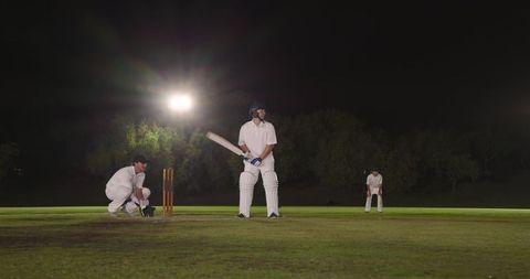 Night cricket batting action at crease under floodlights with wicket keeper and fielder