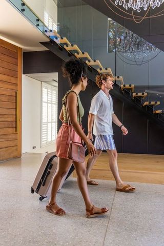 Couple walking into modern foyer pulling suitcase past glass staircase luxury chandelier