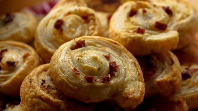 Close-up of golden brown puff pastry pinwheels with savory toppings. Perfect for illustrating recipes, food blogs, bakery menus, or party snack ideas. Highlights texture and color for food photography or culinary websites.