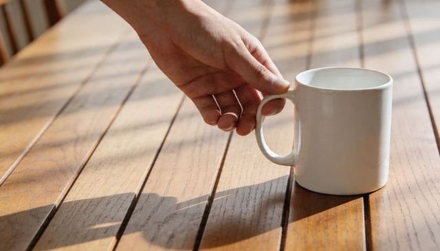 Reaching hand picking up white mug on wooden slat table with sunbeam shadows