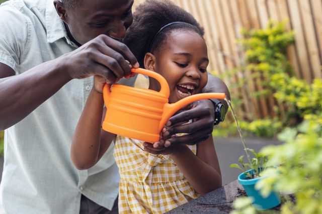 African american father and daughter laughing and watering small potted plants with can in yard. Unaltered, family, togetherness, childhood, gardening and nature concept.