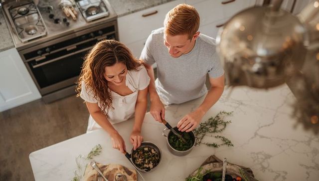 Couple cooking together on marble island chopping herbs and stirring sauce for dinner