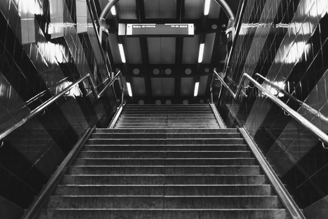The black and white image captures stairs leading up to an underground subway or metro station. The setting is urban, with clean lines and polished surfaces, adding a modern and stylish look. Use this for projects related to city life, transportation, travel, and architectural design.