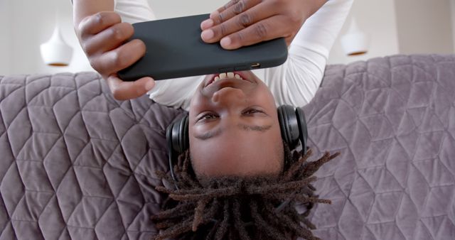 African American boy enjoying time on couch wearing headphones, using smartphone. Ideal for technology, leisure, relaxation, and connectivity themes. Great for lifestyle and tech marketing campaigns.