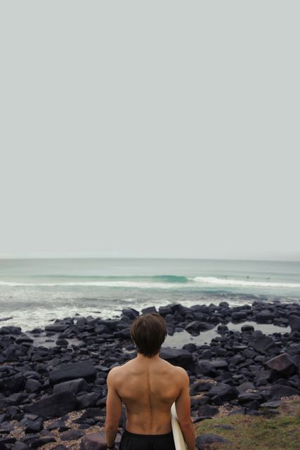 Surfer standing on a rocky shore looking at the ocean waves, holding a surfboard. Great for themes related to adventure, surfing, beach activities, seaside relaxation, nature appreciation, and athletic pursuits. Suitable for travel websites, blogs, magazines, outdoor activity promotions, and surfing events.