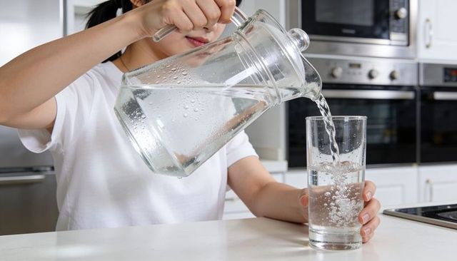 Woman pouring water from glass pitcher into tall glass on modern kitchen counter