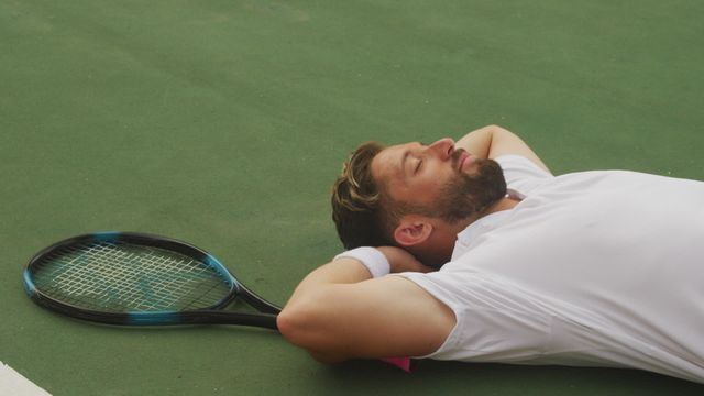 Man playing tennis relaxing on court after strenuous game. Ideal for use in sports apps, fitness websites, magazines detailing athletic recovery principles, tennis tutorials, and adverts promoting sports equipment and activewear.