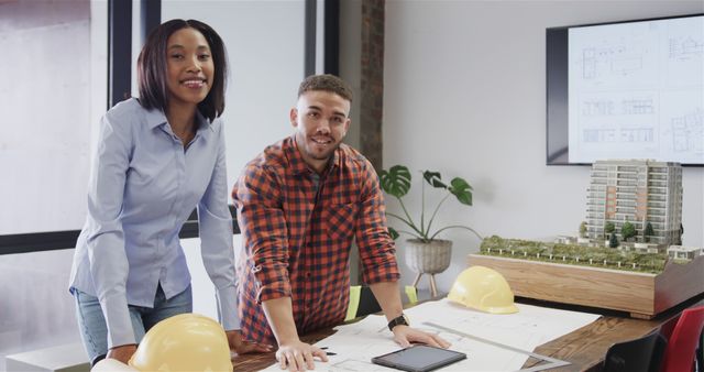 Two architects, a man and a woman, collaborating on a building project in an office. They are leaning over a table with blueprints, hard hats, and an architectural model displayed. The office setting suggests teamwork and professional planning, suitable for use in articles or promotions about architectural firms, construction projects, teamwork in engineering, and office collaboration.