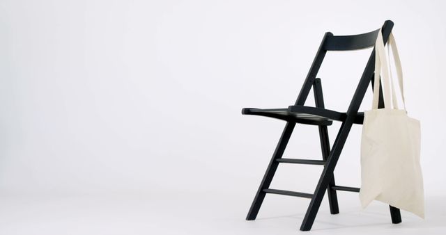 A black folding chair with a canvas tote bag hanging on it, against a white background with copy space. The simplicity of the setup suggests a minimalist or contemporary aesthetic.