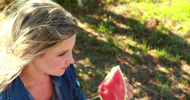 Young woman enjoying eating watermelon outdoors on a sunny day. Ideal for themes related to healthy eating, summer activities, fresh produce, outdoor enjoyment, and lifestyle. Captures a casual, cheerful, and healthy moment in natural light, suitable for use in advertisements, blogs, magazines, and social media posts promoting wellness, summertime, or diets.