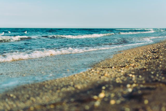 This stock photo features a serene sandy beach with gentle waves lapping against the shore and a clear blue sky above. Ideal for travel blogs, vacation marketing materials, relaxation and wellness themes, and nature-related publications to evoke a sense of peace and tranquility.