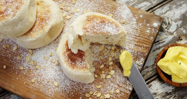 Freshly baked English muffins are sprinkled with flour on a wooden board, one split open revealing a jam filling. A knife and a small basket with butter add to the cozy breakfast atmosphere, inviting a delightful start to the day.