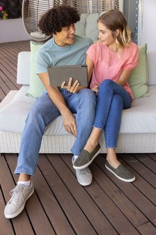 Young couple sharing tablet on patio wicker sofa, relaxing and chatting on wooden deck