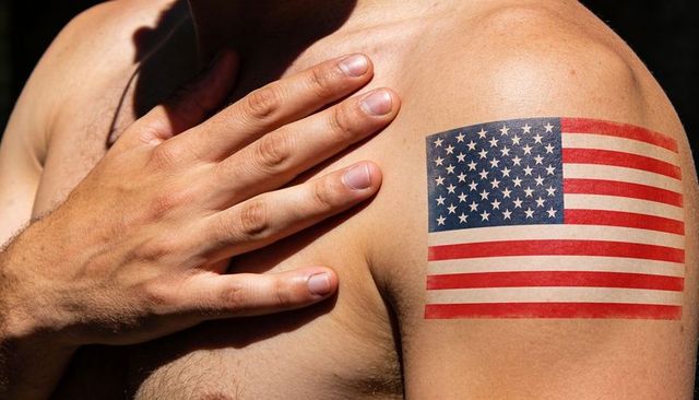 Man placing hand on chest showing american flag tattoo on upper arm in sunlight