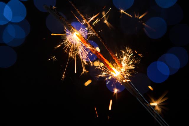 Sparkler igniting with bright sparks against a dark background with defocused blue lights. Ideal for use in holiday and celebration themes, such as New Year and Christmas. Perfect for festive greeting cards, party invitations, and promotional materials for events.