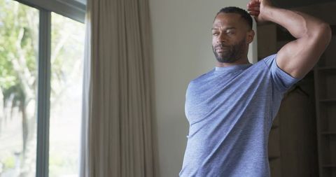 Mature man stretching by window wearing light-blue t-shirt for morning stretch and calm