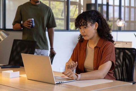 Hispanic woman checking smartwatch while typing on laptop in modern collaborative office