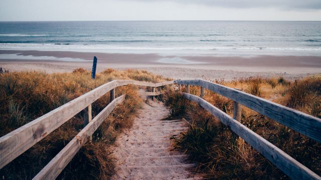 Wooden pathway leading to serene beach with calm ocean waves on a cloudy day. Ideal for travel blogs, vacation planning websites, and coastal living articles to depict tranquil beach scenes and seaside relaxation. Perfect for promoting summer trips and surfing spots.