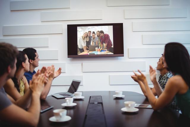 Business professionals are seen applauding during a video conference in a modern conference room. This image can be used for articles or presentations about remote work, virtual meetings, corporate teamwork, and modern office environments.