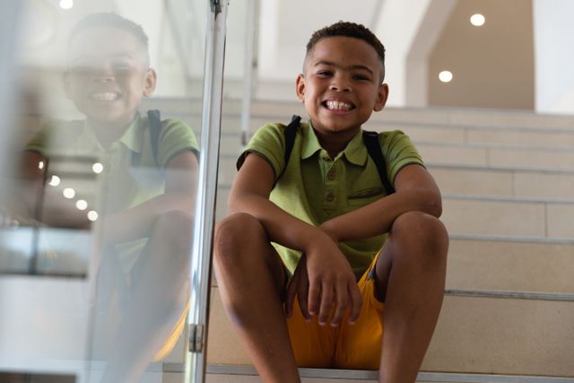 Young African American boy sitting on steps in a school building, smiling at the camera. He is wearing a green polo shirt and yellow shorts, with a backpack on his shoulders. This image is ideal for educational materials, back-to-school promotions, and childhood development articles.