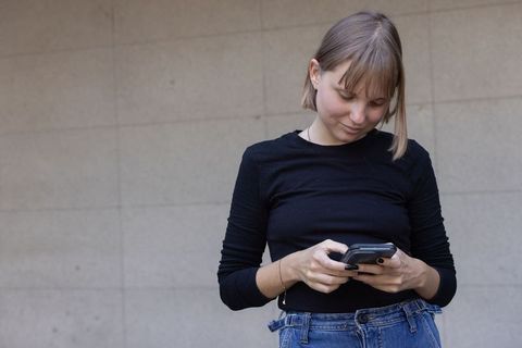Young woman texting on smartphone by gray urban wall wearing black top and high-rise jeans