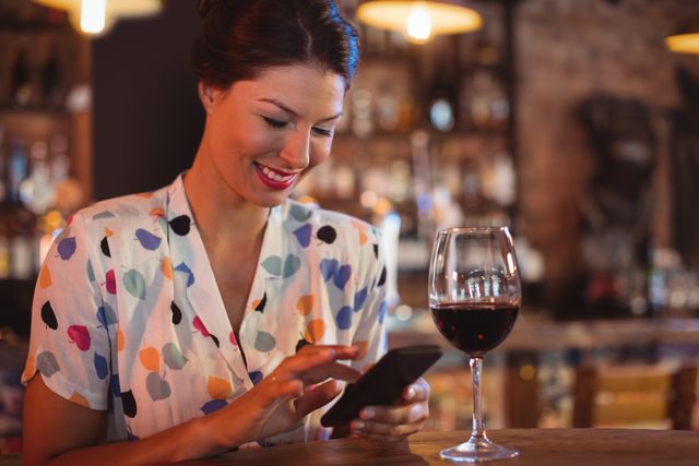 Young woman smiling while using mobile phone in a pub, enjoying a glass of red wine. Ideal for themes related to social media, nightlife, relaxation, leisure activities, and modern lifestyle.