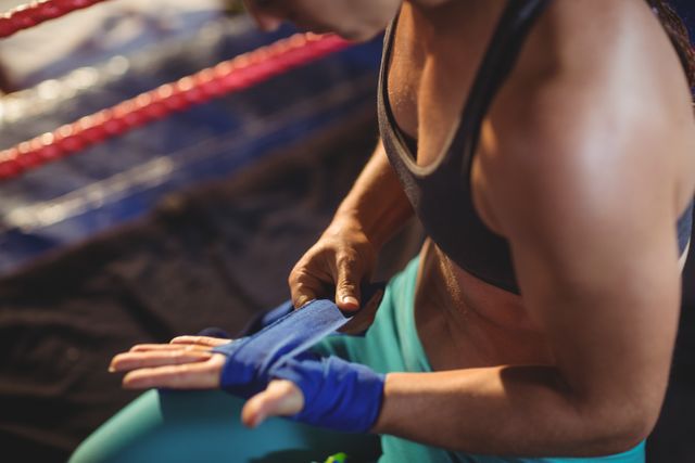 Female boxer wrapping hands with blue strap in boxing ring. Ideal for use in sports and fitness articles, training programs, motivational content, and advertisements for athletic gear. Highlights themes of strength, preparation, and focus.