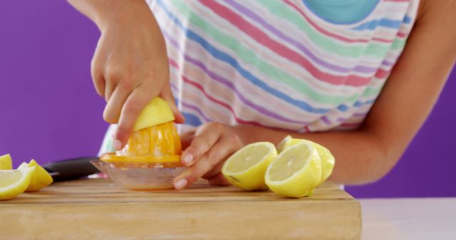 Woman juicing lemons in vibrant kitchen, wearing colorful striped top, preparing fresh citrus juice on wooden cutting board. Ideal for illustrating concepts of healthy living, summer recipes, fresh ingredients, and kitchen activities.