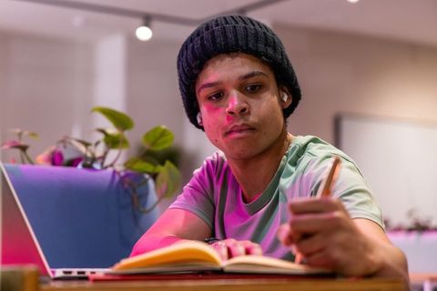Teen student writing in notebook while studying with laptop and beanie indoors