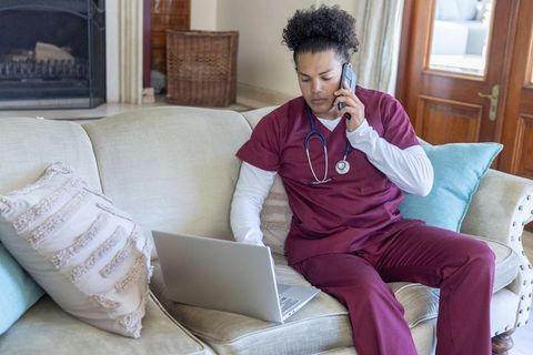 Male nurse working remotely from home on couch, speaking on phone while using laptop