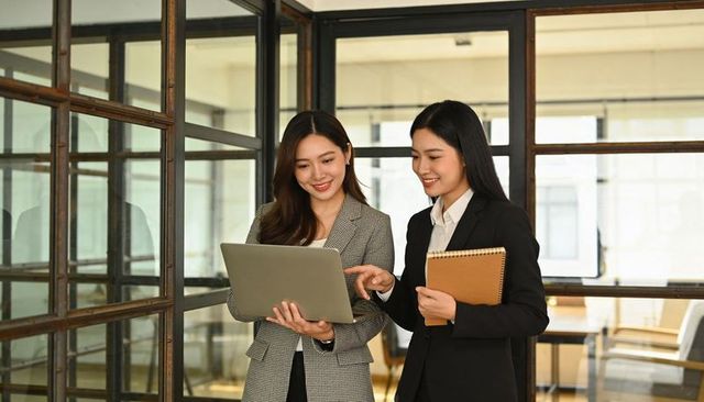 Two professional women collaborating over laptop in modern glass office, business teamwork