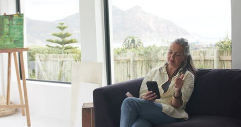 Mature woman relaxing on sofa checking smartphone and gesturing near easel by big window
