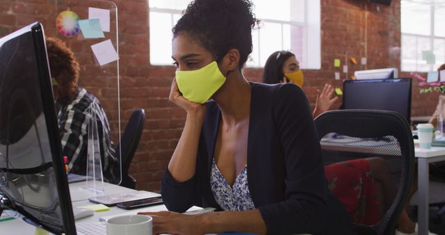 Biracial businesswoman having image chat sitting in front of computer wearing face mask - Download Free Stock Photos Pikwizard.com