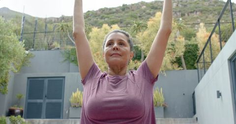 Mature woman raising arms during outdoor stretch in modern backyard garden
