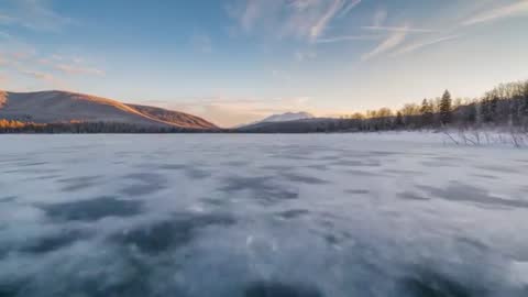 Drone Pulling Back Over Frozen Alpine Lake Revealing Textured Ice and Sunlit Ridge at Sunrise
