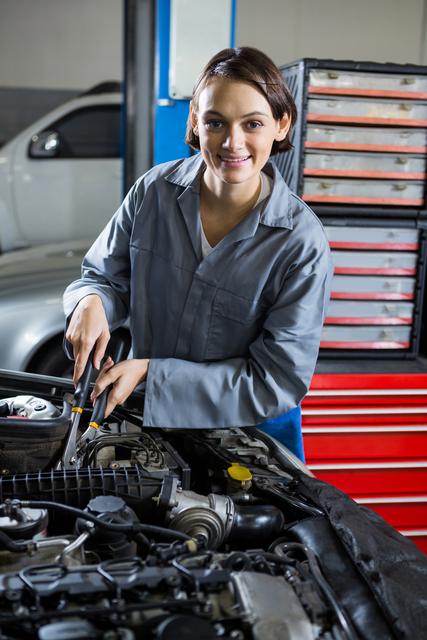 A smiling female mechanic is repairing a car in an auto repair garage. She is working on the engine, using tools, and wearing a uniform. This image can be used to represent women in traditionally male-dominated professions, automotive service, or mechanical expertise. Ideal for articles, advertisements, or promotional materials related to mechanics, car repair services, and gender diversity in the workplace.